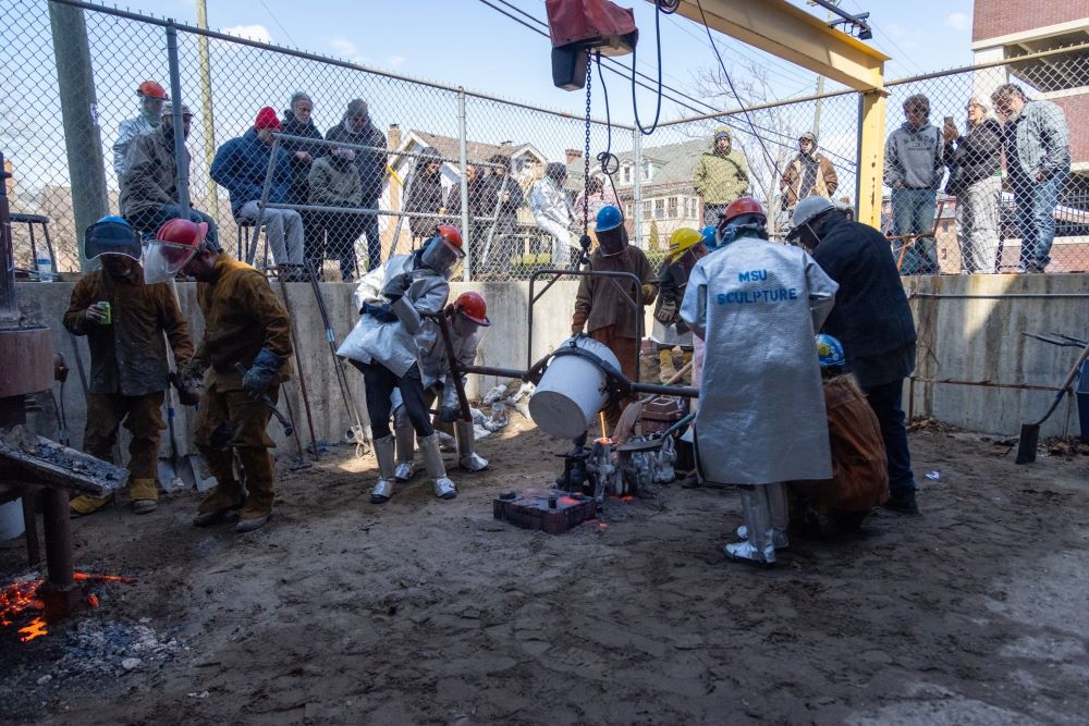 People in protective jackets, gloves, and helmets work together pour molten metal into molds in an industrial setting outside. At the street level are people behind a chain-length fence watching the iron pour.