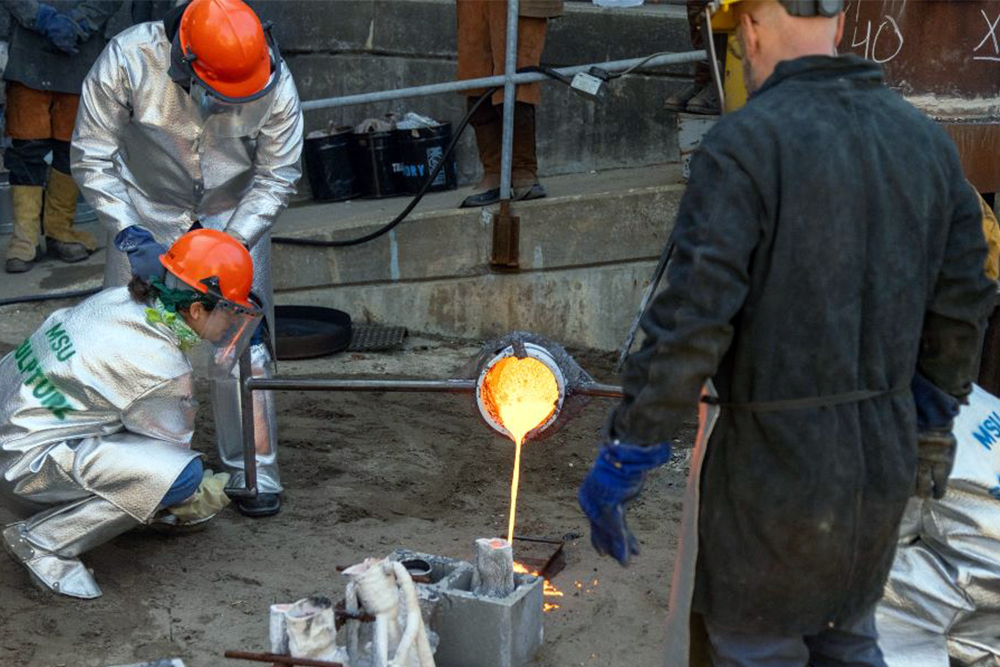 People in protective jackets, gloves, and helmets work together pour molten metal into molds in an industrial setting outside.