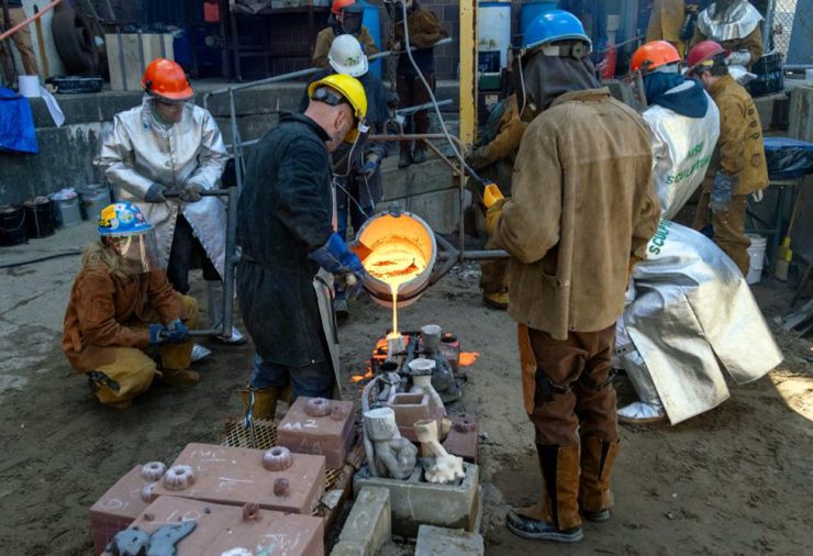 People in protective jackets, gloves, and helmets work together to pour molten metal into molds in an industrial setting outside.