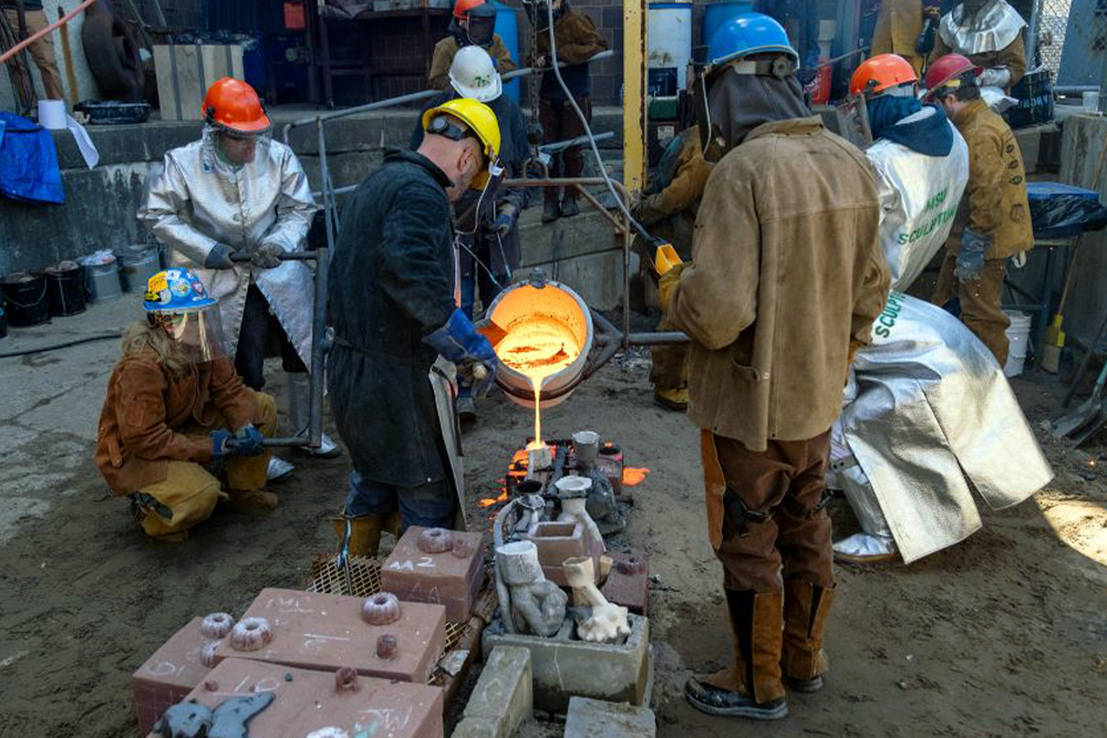People in protective jackets, gloves, and helmets work together to pour molten metal into molds in an industrial setting outside.