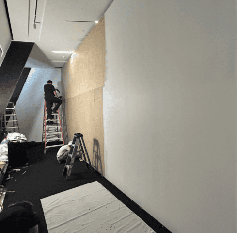 Two men attach beige material to a large white wall. One stands on a ladder while the other crouches down next to a smaller ladder.