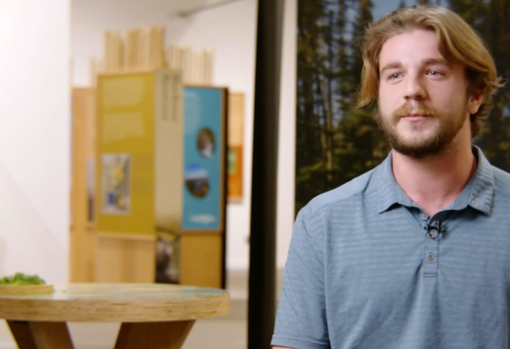 A man wearing a blue collared shirt sits in a museum environment.