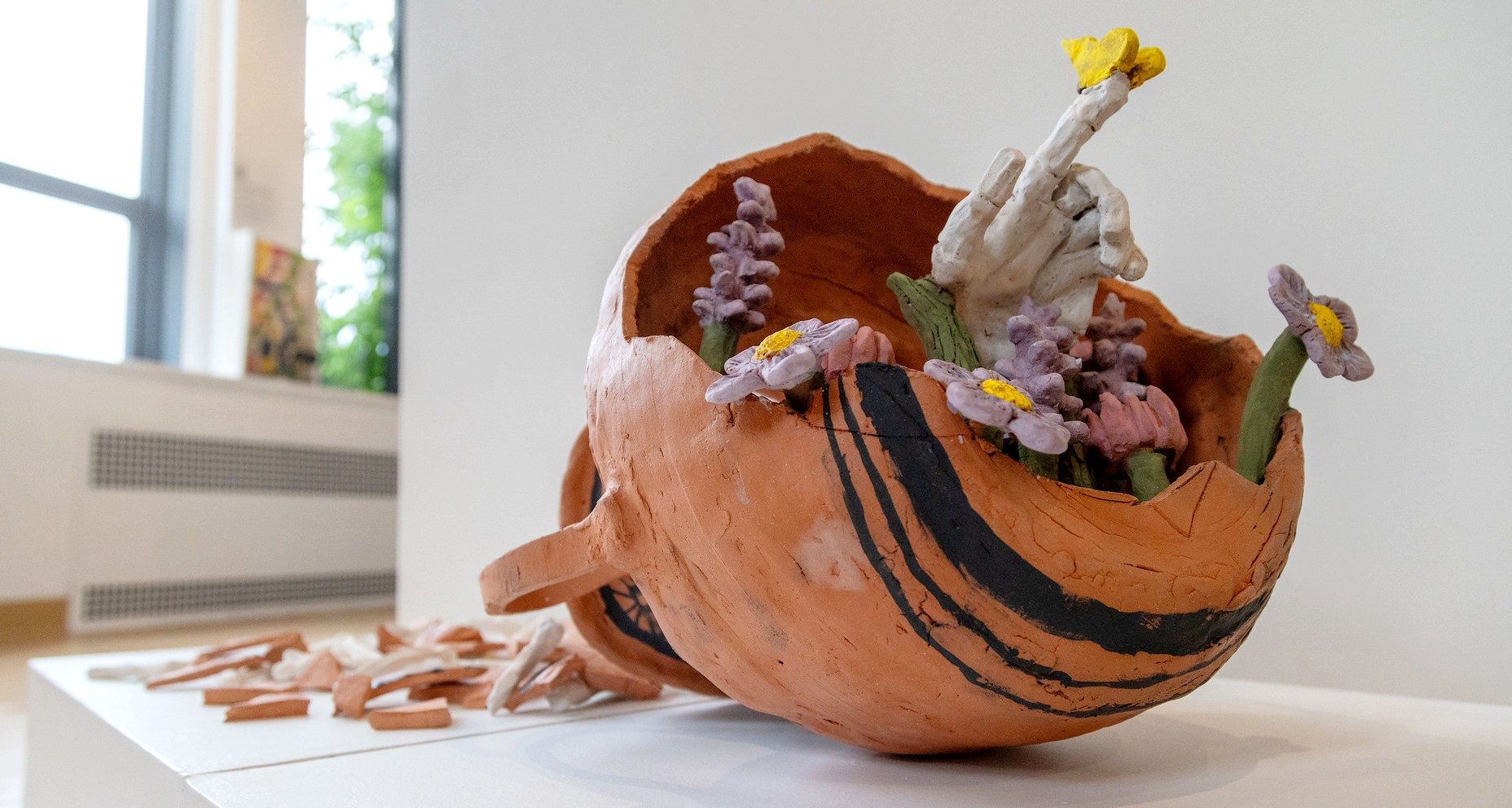Ceramic sculpture resembling a broken terracotta vessel filled with colorful flowers and sculpted hands.