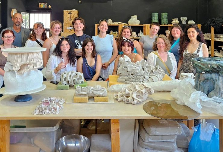 Students and instructors from the Ceramic Arts in Greece education abroad program pose together in a ceramics studio behind a table filled with sculptural ceramic works.