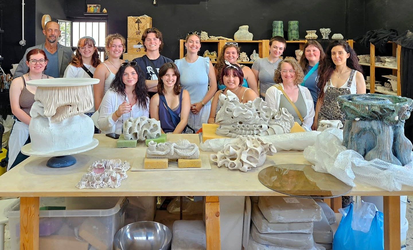 Students and instructors from the Ceramic Arts in Greece education abroad program pose together in a ceramics studio behind a table filled with sculptural ceramic works.