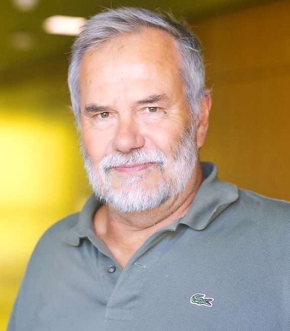 Portrait of David Stowe, a man with gray hair and a beard wearing a gray polo shirt, standing a warmly lit indoor space.