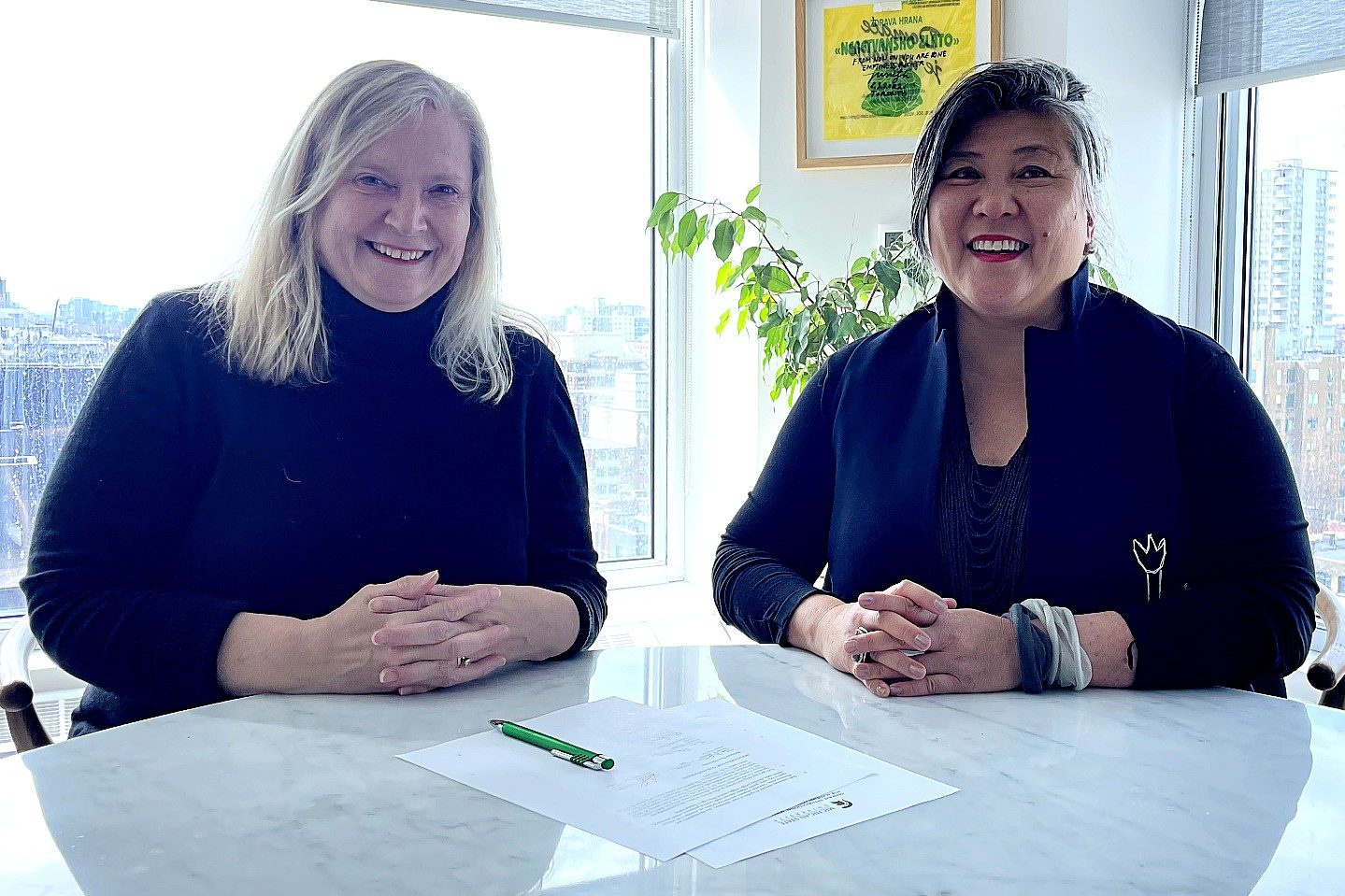Two women smile while seated together at a table in a bright office, with papers and a green pen in front of them and city buildings visible through the windows behind them.