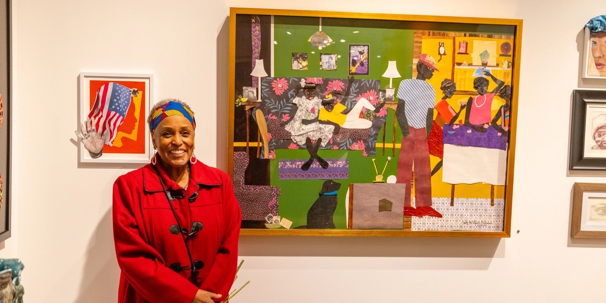 Judy Bowman wearing a red coat and colorful headwrap, holds flowers and smiles beside her large collage depicting a family gathered in a living room.