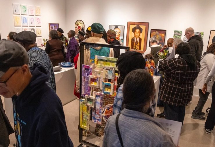 A crowd moves through a gallery filled with portraits, collages and mixed-media artwork displayed on the walls and freestanding panels.