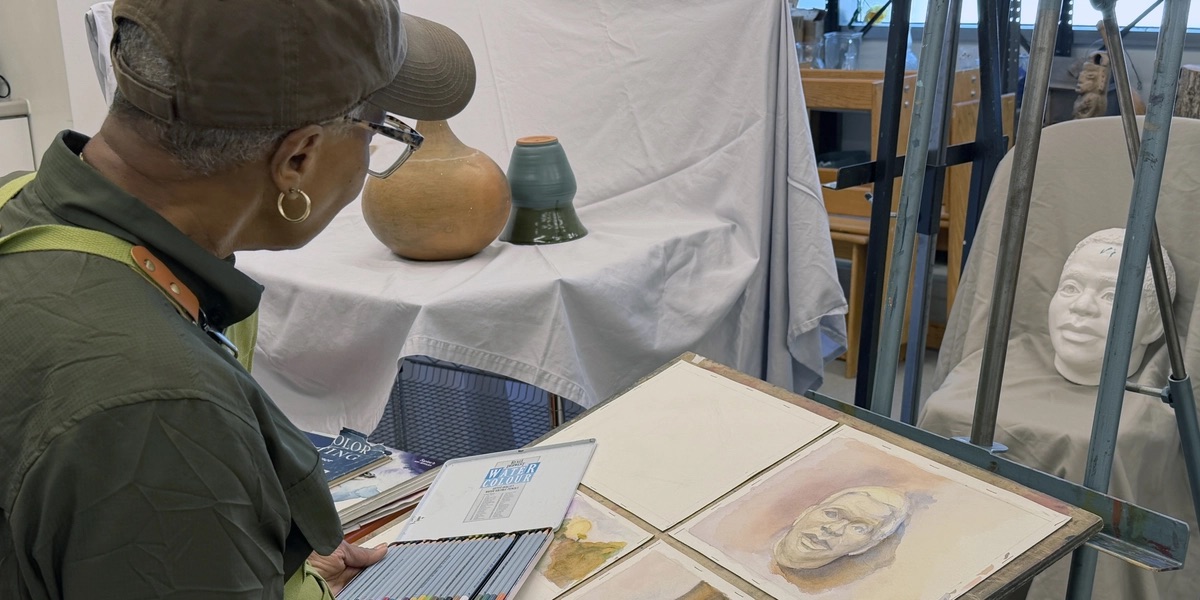 A SOLHOT 60+ participant wearing a cap and green apron works at a table covered with watercolor paintings and art supplies. Ceramic vessels and a sculpted head serve as a reference nearby.