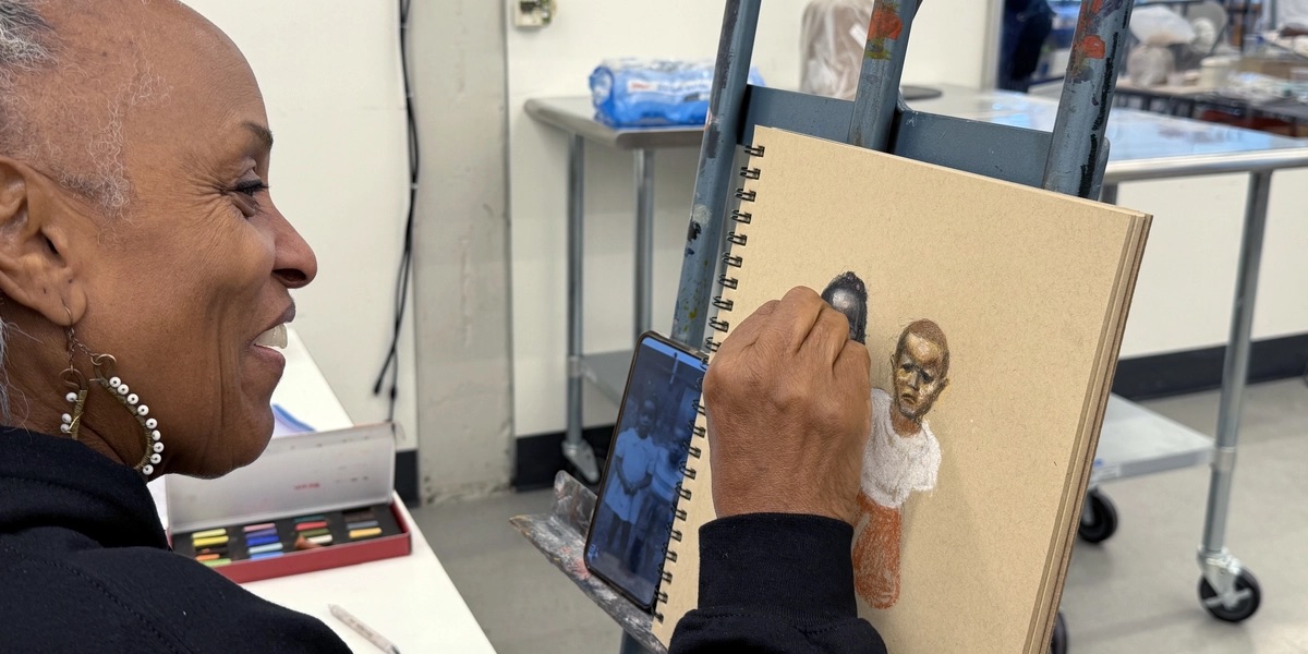 Judy Bowman smiles as she uses pastels to draw two people on tan paper displayed on an easel. A cellphone beside the drawing displays her reference image.