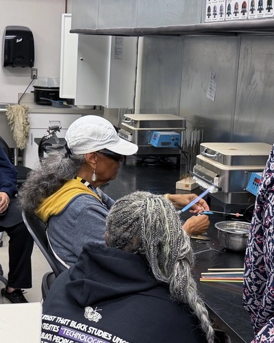 A participant wearing a navy SOLHOT 60+ hoodie watches as Dr. S.A. Simon shapes glass over a flame at a beadmaking station.