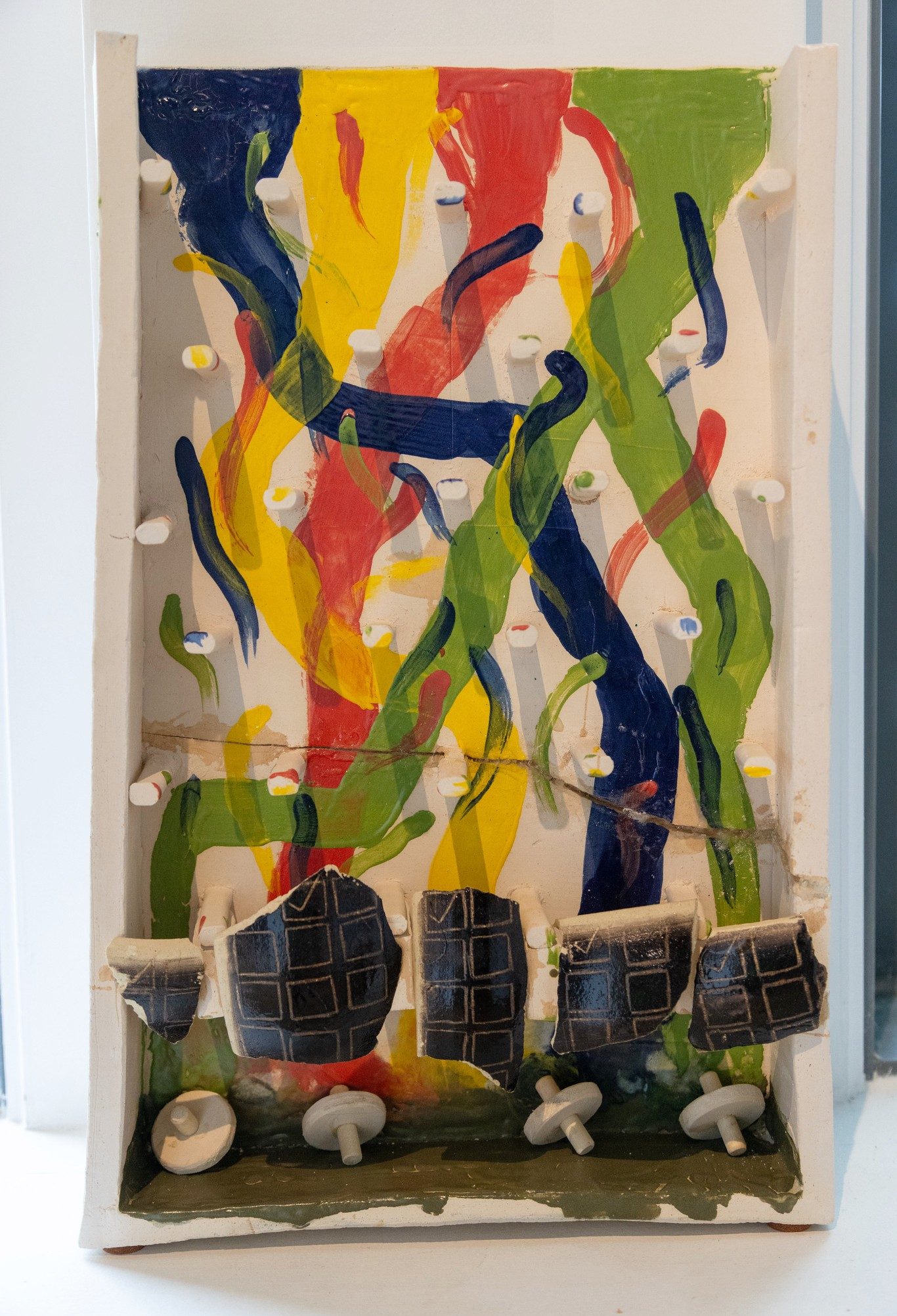 Mixed-media ceramic artwork with sweeping red, yellow, green, and blue brushstrokes, protruding pegs, and dark geometric forms.