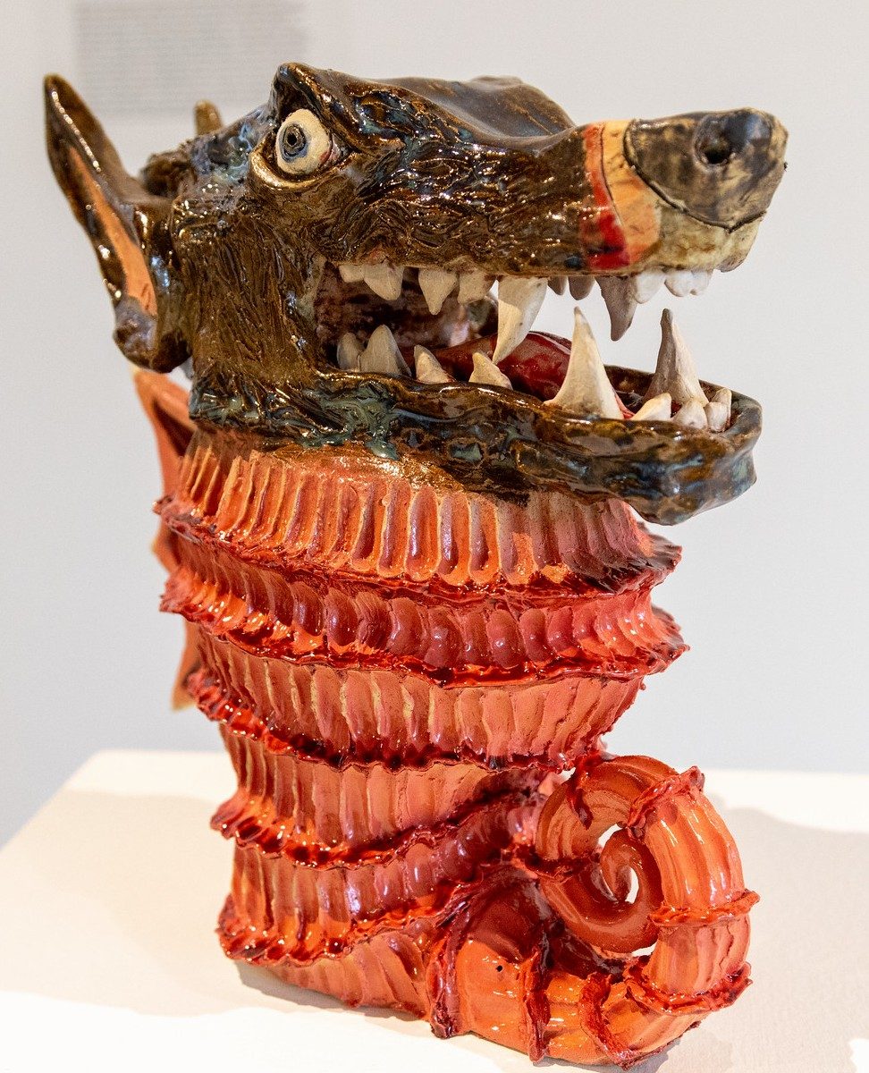 Glazed ceramic sculpture featuring a wolf-like head with an open mouth and prominent teeth atop a textured red-orange form.
