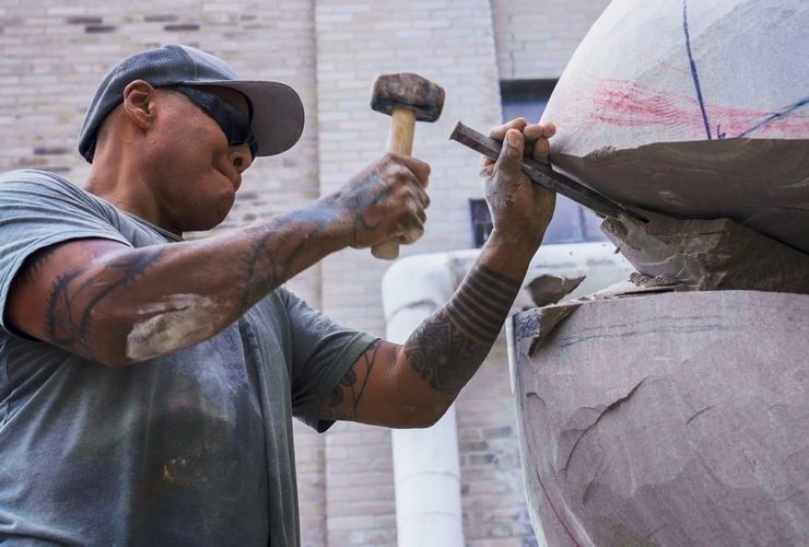 Sculptor Jason Quigno uses a hammer and chisel to carve a stone sculpture.