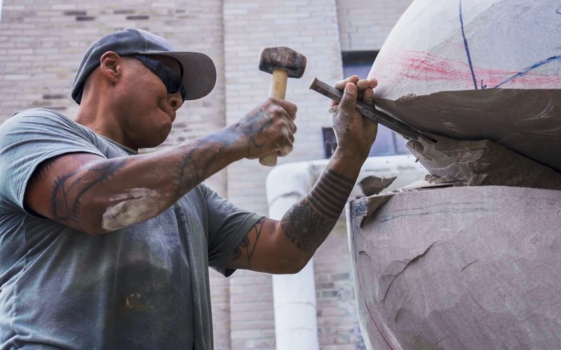 Sculptor Jason Quigno uses a hammer and chisel to carve a stone sculpture.