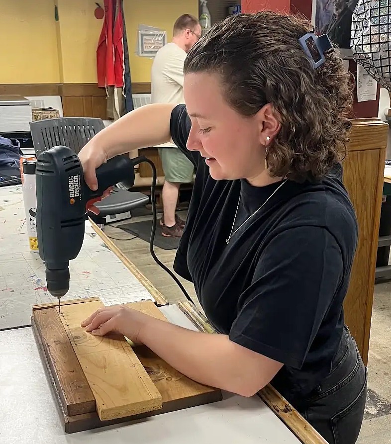 A student uses a power drill to make holes in a wooden board while working on a object in a studio.