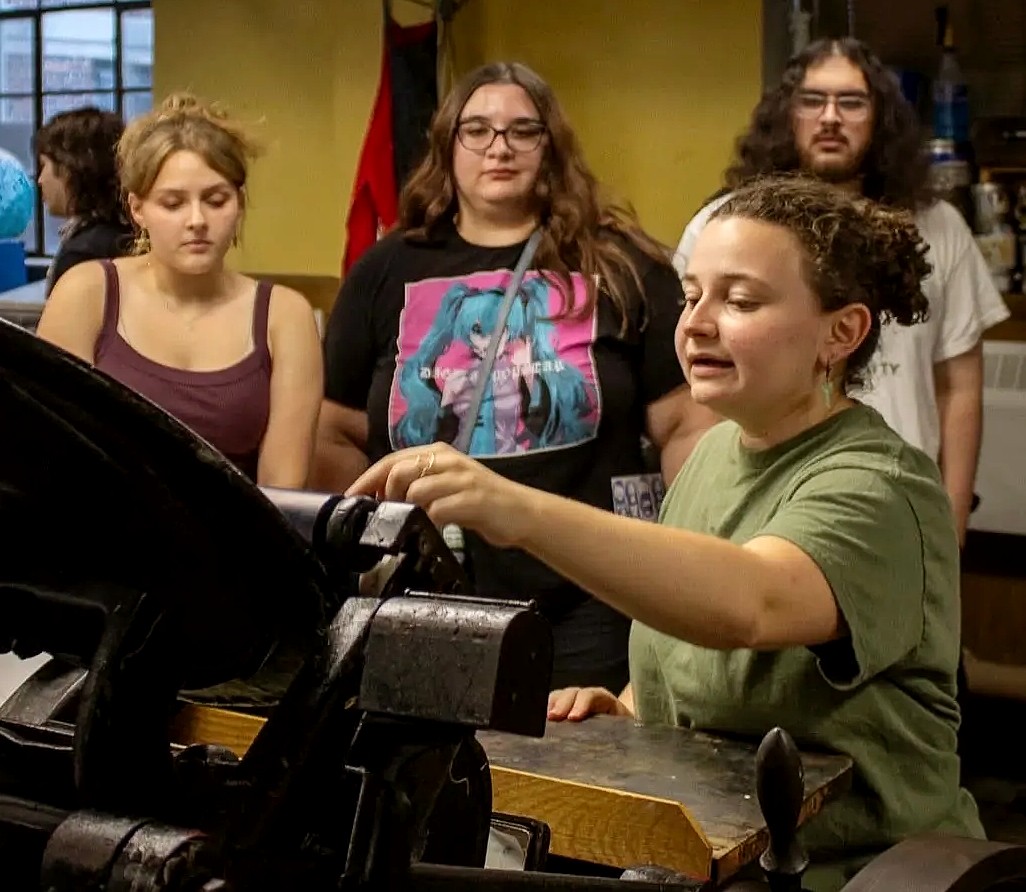 A group of student gathers around a historic printing press in a printmaking workshop. One student operates the press while the others observe and learn about the printing process.