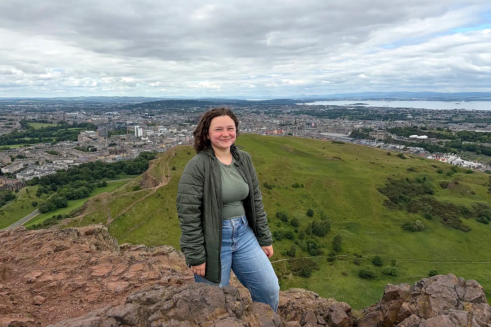 A person standing on a rocky overlook with a view of Edinburgh in the background.