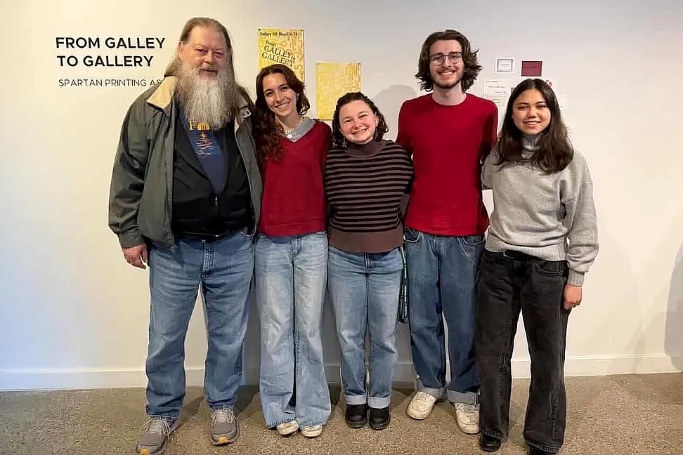 A group of five people, including an older man with a long white beard and four young adults, stand together smiling in front of an exhibition wall titled 