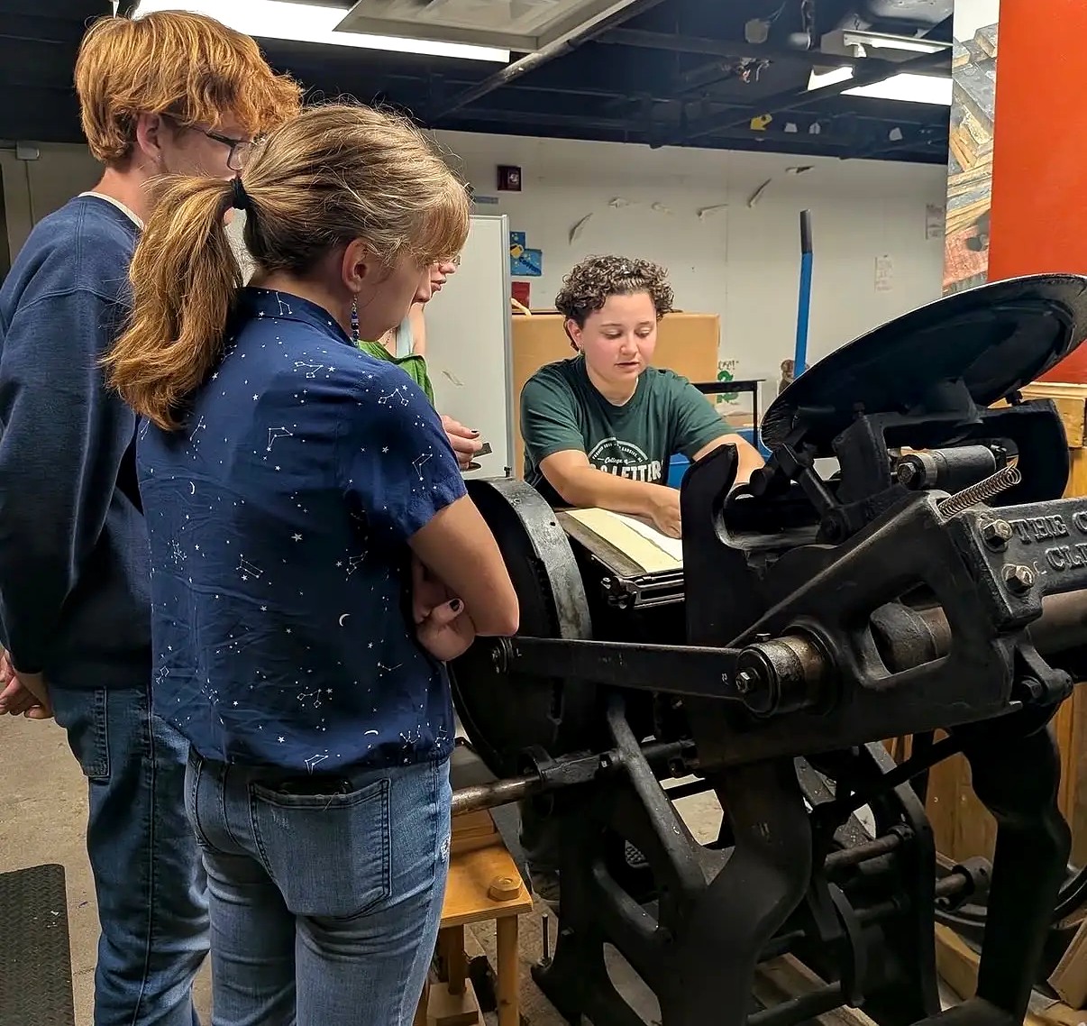 A group of students gathers around a historic printing press in a printmaking workshop. One student operates the press while the others observe and learn about the printing process.
