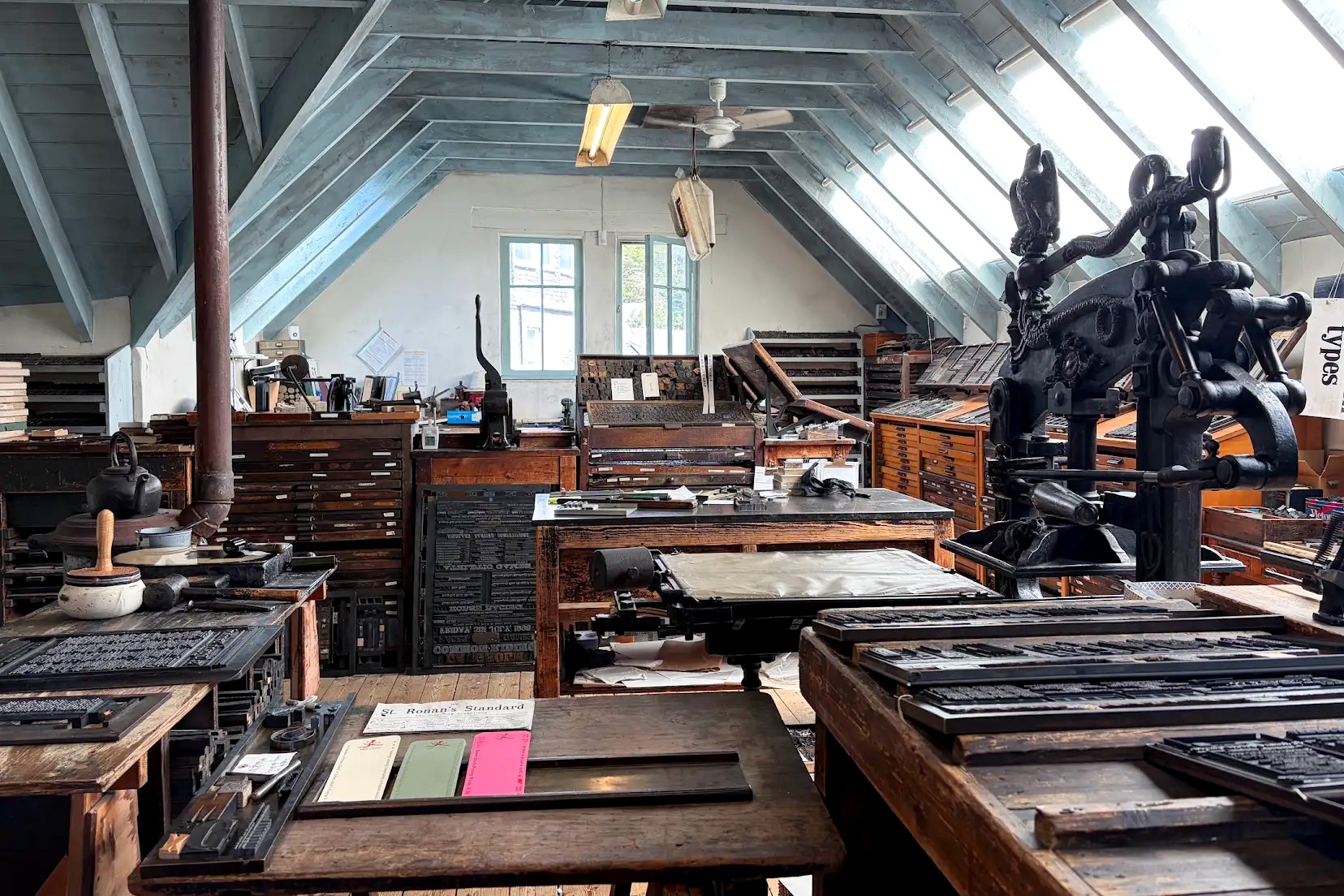 A historic letterpress printing workshop filled with antique printing presses, wooden type cabinets, trays of metal type, and worktables. Natural light streams through large slanted skylights, illuminating the traditional printmaking equipment and tools.